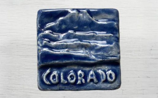 Ceramic tile depicting a mountain landscape in blue tones, labeled "COLORADO," showcasing local artistry and geographical significance.