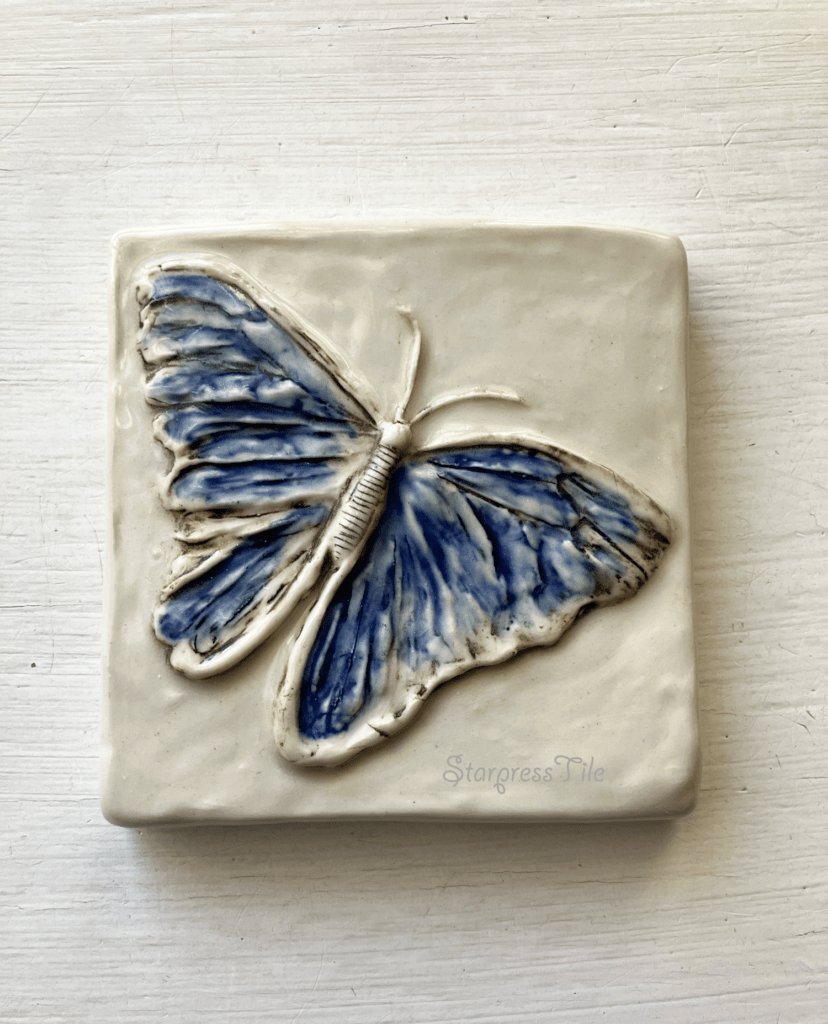Blue butterfly relief ceramic tile with intricate detailing, featuring a textured white background. Perfect for decor or artistic display.