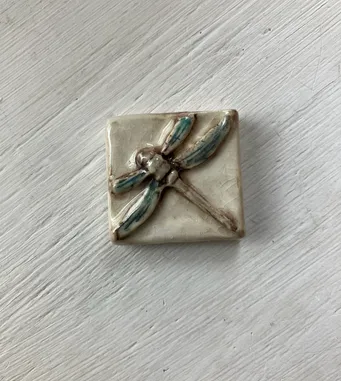 Ceramic tile featuring a dragonfly design in soft blues and browns, highlighting intricate craftsmanship. Ideal for home decor or art discussion.