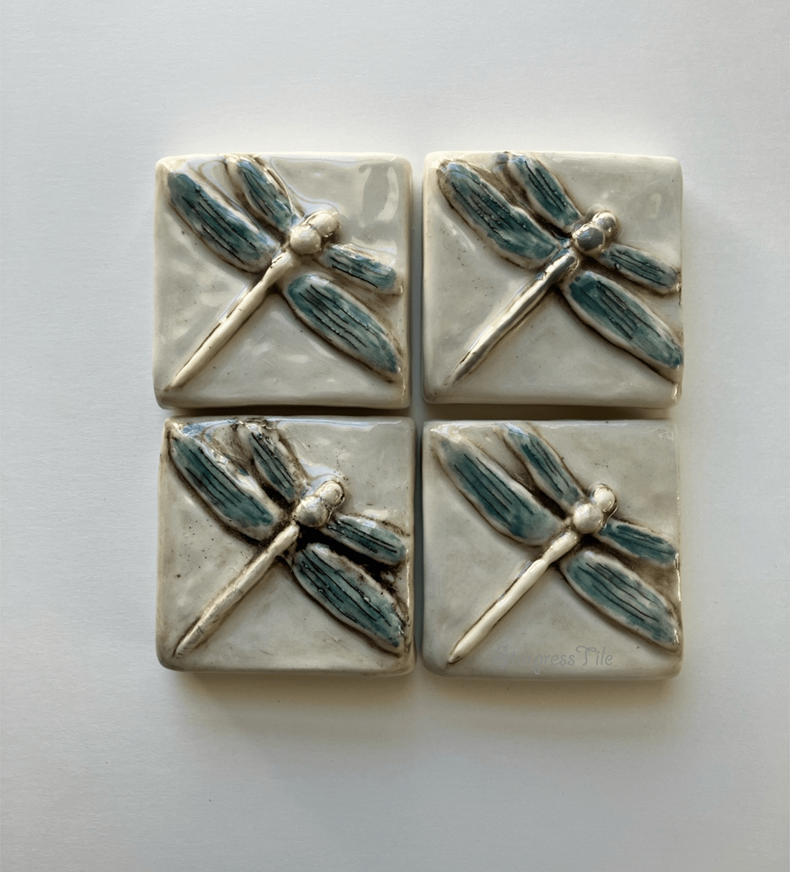 Four ceramic tiles featuring embossed dragonfly designs in a watercolor hand painted teal and brown highlight. The artwork adds a natural element to home decor.